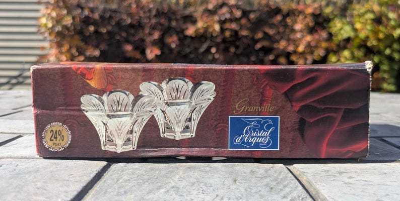 May include: A rectangular box with a maroon and red design featuring two clear crystal candle holders. The box has the text "Granville" and a blue label with "Cristal d'Arques". A gold seal reads "24% PBO".