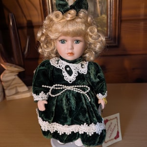 May include: A porcelain doll with blonde curly hair and blue eyes. The doll wears a dark green velvet dress with white lace trim and a matching bow. A tag reading "Certificate of Authenticity" is attached.