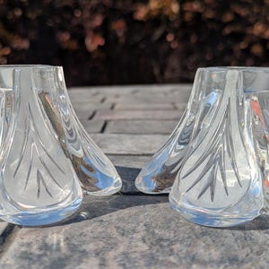May include: Two clear glass candle holders with a petal-like design. The candle holders are sitting on a gray surface. The glass has a reflective quality, and the design is etched into the glass.