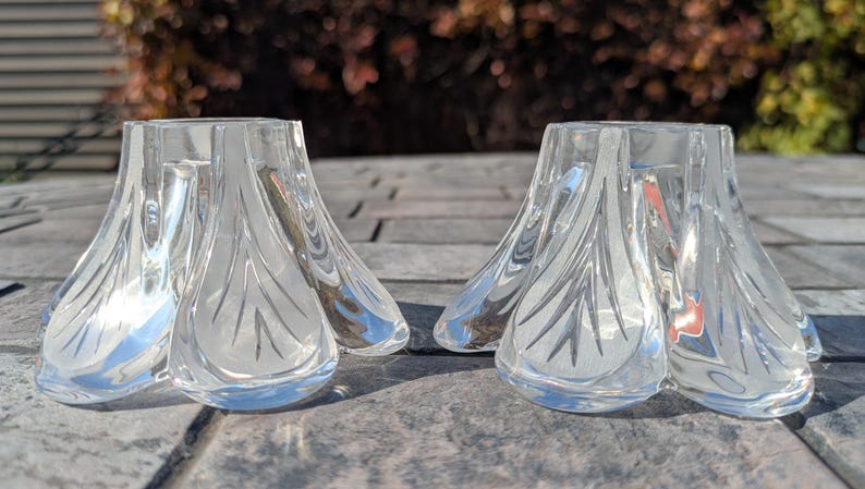 May include: Two clear glass candle holders with a petal-like design. The candle holders are sitting on a gray surface. The glass has a reflective quality, and the design is etched into the glass.