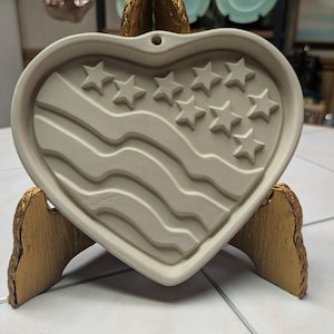 May include: A ceramic heart-shaped cookie cutter with a raised design of the American flag. The cutter is light brown and has a hole at the top for hanging.