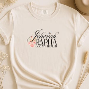 May include: A cream-colored short-sleeve t-shirt featuring the black text "Jehovah RAPHA GOD MY HEALER" printed on the chest. The design includes a small illustration of a bee and a cluster of pink roses near the lettering.