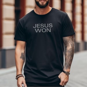 May include: Black t-shirt with the text "JESUS WON" in gray block letters. The short-sleeved shirt is made of a soft material. The shirt is a casual wear item.
