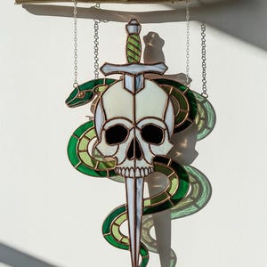 May include: Stained glass artwork depicting a skull impaled by a sword, with a green snake coiled around it. The skull is white with black eye sockets, and the sword has a green handle. The snake is green and white. It hangs from chains.