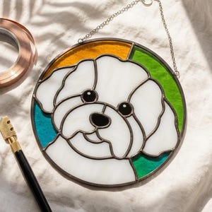 May include: A round stained glass window featuring a white dog face with black eyes and nose, framed by a green, yellow, and blue border. A roll of copper foil tape and a glass cutter are also in the image.