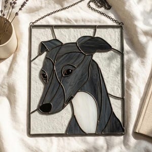 Puede incluir: Un panel rectangular de vidriera que muestra un perro de color gris oscuro y negro con el pecho blanco, sobre un fondo texturizado de color blanco roto. La pieza está enmarcada en metal oscuro e incluye una cadena de metal negro para colgar.