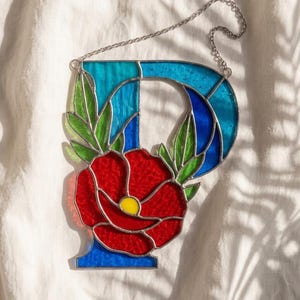 May include: A stained glass letter P suncatcher featuring shades of blue and turquoise glass. A large red flower with a yellow center and green leaves is integrated into the design. The piece includes a silver metal chain for hanging purposes.