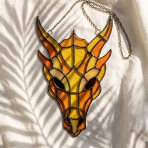 Op de afbeelding: Een glas-in-lood zonnevanger in de vorm van een drakenkop met levendige gele en oranje glassegmenten, verbonden door donkere metalen soldeerlijnen. Het object is voorzien van een dunne metalen ketting aan de bovenzijde om het op te kunnen hangen.