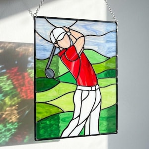 Può includere: Un pannello rettangolare in vetro colorato che raffigura un golfista con camicia rossa, cappellino bianco e pantaloni bianchi durante uno swing. Lo sfondo presenta colline verdi ondulate e un cielo azzurro, il tutto unito da linee di piombo.