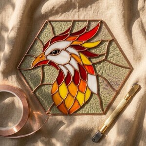 May include: A stained glass phoenix in a hexagonal frame, with red, orange, yellow, and white glass pieces. The bird's head has an orange beak and eye. Copper foil and a glass cutter are next to the artwork.