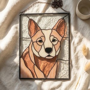 May include: A rectangular stained glass panel featuring a dog with tan and cream-coloured glass pieces. The dog has pointed ears and a dark nose, framed in a thin black metal border with a metal chain attached to the top for hanging purposes.