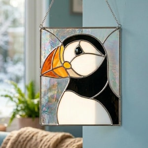 May include: A rectangular stained glass suncatcher featuring a puffin with a black and white body and a bright orange beak. The bird is set against an iridescent, textured glass background, framed in metal, and suspended by a silver chain.
