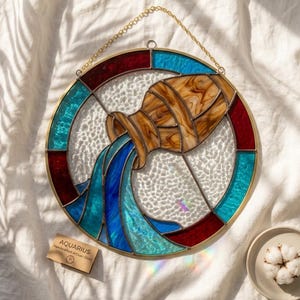 Op de afbeelding: Een ronde glas-in-lood zonnevanger met het sterrenbeeld Waterman, met een bruine kruik die blauw water giet. Het ontwerp is gevat in goudkleurig metaal met rode en blauwe randsegmenten en een achtergrond van getextureerd, transparant glas.