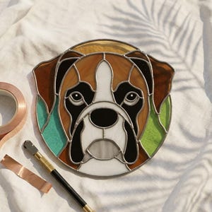 May include: A stained glass artwork depicting a Boxer dog's face, featuring brown, black, white, and green glass pieces. The artwork is surrounded by a metal frame. A roll of copper tape and a glass cutter are next to the artwork.