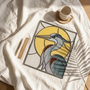 May include: A rectangular stained glass panel featuring a blue and brown heron silhouette against a large yellow circular sun. The panel is positioned next to a gold pen and a wooden ruler measuring 15 cm in length, with a white ceramic mug nearby.