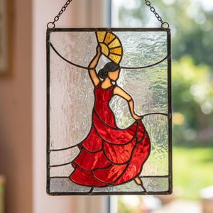 May include: A rectangular stained glass suncatcher featuring a figure in a red dress holding a yellow fan. The figure is depicted in a dance pose against a textured clear glass background, all framed in dark metal with a hanging chain.