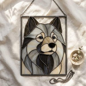 May include: A rectangular stained glass panel featuring a stylised portrait of a dog with pointed ears. The piece is crafted from various shades of cream, grey, and black glass pieces joined by dark metal solder lines, suspended by a metal chain.