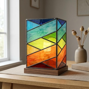 May include: A rectangular stained glass table lamp featuring a vibrant rainbow color gradient from blue at the top to red at the bottom. The glass panels are held together by dark metal soldering and mounted on a solid rectangular wooden base.