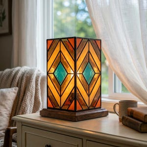 Könnte beinhalten: Eine rechteckige Tiffany-Tischleuchte mit einem geometrischen Muster aus bernsteinfarbenen, orangefarbenen und blaugrünen Glaspaneelen, die von einem dunklen Metallrahmen gehalten werden. Die Leuchte steht auf einem quadratischen Holzsockel.