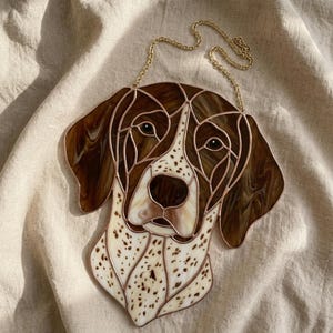 Può includere: Ritratto di cane in vetro colorato, un Bracco tedesco. L'opera presenta una testa di cane macchiata marrone e bianca, con una catena dorata per appenderla. I pezzi di vetro sono delineati in rame.
