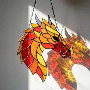 Op de afbeelding: Een glas-in-lood zonnevanger in de vorm van een drakenkop met levendige rode, oranje en gele glazen segmenten, omlijnd met donker metaalsoldeer. De draak heeft twee kleine grijze hoorns en hangt aan een dunne zwarte metalen ketting.