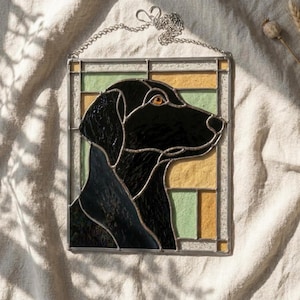 Può includere: Un pannello rettangolare in vetro colorato raffigurante il profilo di un cane nero con un occhio color ambra. Il cane è posto su uno sfondo di pannelli di vetro geometrici in tonalità verde pallido, giallo e vetro trasparente testurizzato, uniti da profili metallici argentati.