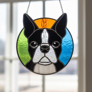 May include: Stained glass Boston Terrier dog suncatcher with black, white, orange, green, and blue colors. The dog's face is centered in a circular design, hanging from a chain. The dog's face is black and white.