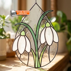 May include: A teardrop-shaped stained glass suncatcher featuring two white snowdrop flowers with green stems and leaves, outlined in dark metal solder. The piece includes a small rectangular label with the text SVG CUTTING MACHINE COMPATIBLE.