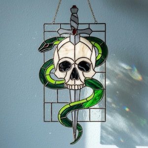 Könnte beinhalten: Ein Buntglas-Suncatcher mit einem weißen Totenkopf, der auf einem vertikalen silberfarbenen Dolch zentriert ist und von einer leuchtend grünen Schlange umwunden wird. Das Design ist in einem rechteckigen Gitter aus Klarglas eingefasst.