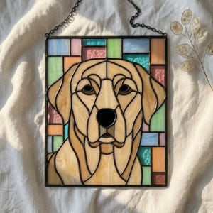 Op de afbeelding: Een rechthoekig glas-in-lood paneel met een portret van een gele Labrador retriever. De hond is gemaakt van crème- en beigegekleurde glasstukken met donkere soldeerlijnen. De achtergrond bestaat uit een raster van pastelkleurige glasvierkanten.