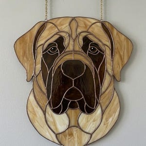 May include: Stained glass artwork depicting a dog's head, featuring shades of brown and tan. The piece is designed with intricate lines and details, showcasing the dog's face. It hangs from two gold-colored hooks.