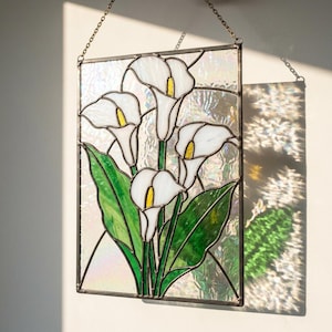 May include: Stained glass panel featuring calla lilies. The artwork showcases white flowers with yellow centers and green leaves, set against an iridescent background. The panel is framed in a dark metal and hangs from a chain.