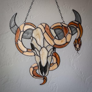 Peut inclure: Un attrape-soleil en vitrail représentant un crâne de bélier crème au centre avec des cornes grises courbées. Un long serpent sinueux aux segments orange, marron et crème s'enroule autour du crâne, avec des yeux rouges et une chaîne.