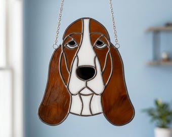 May include: A stained glass suncatcher shaped like a Basset Hound dog head, featuring brown and white glass segments joined by dark metal solder lines. The piece includes a silver-toned metal chain attached to the top for hanging display purposes.