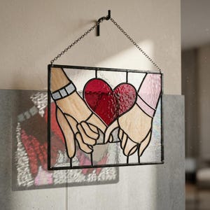 May include: A rectangular stained glass panel featuring two hands with fingers intertwined, centred around a large, textured deep red heart. The piece is framed in black metal and suspended by a silver-toned metal chain from a wall hook.