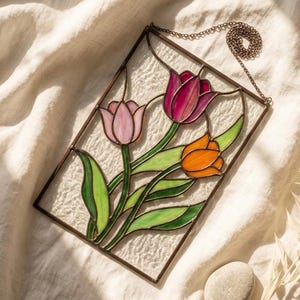 May include: A rectangular stained glass panel depicting three tulips in pink, orange, and purple, with green leaves. The flowers are set against a textured white background within a brown frame, with a hanging chain.