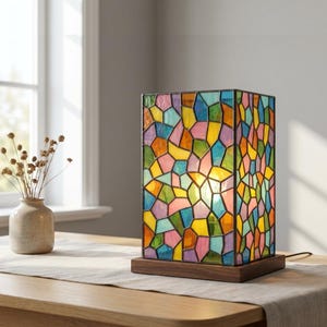 Op de afbeelding: Een rechthoekige tafellamp met een glas-in-lood kap met een veelkleurig mozaïekpatroon van geometrische vormen in blauw, geel, roze, groen en oranje, geplaatst op een donkere houten voet met een zichtbaar netsnoer.