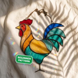 Könnte beinhalten: Ein Buntglas-Sonnenfänger in Form eines Hahns mit rotem Kamm und Kehllappen, gelbem Kopf und Körper sowie einem Schwanz aus blauen und türkisfarbenen Segmenten. Das Stück hängt an einer Metallkette und trägt ein grünes Etikett mit dem Text BEGINNER PATTERN.