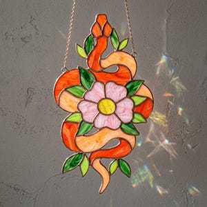 May include: A stained glass suncatcher featuring an orange snake coiled around a central pink flower with a yellow centre, accented by green leaves. The piece is suspended by a thin metal chain and displays vibrant, multi-coloured glass segments.