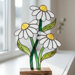 May include: A stained glass sculpture of three daisies. The white petals are outlined in black and have yellow centres. The green stems and leaves are attached to a wooden base. The sculpture is displayed on a white windowsill.