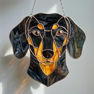 Peut inclure: Tête de chien teckel en vitrail avec un cadre cuivré. Le visage du chien présente du verre noir et jaune, avec une chaîne pour l'accrocher. Le verre a un effet marbré et les yeux du chien sont bruns.