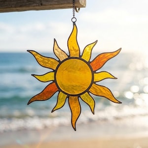 May include: A sun-shaped stained glass suncatcher featuring a central yellow circular piece surrounded by alternating wavy yellow and orange glass rays, all held together by dark metal soldering and suspended by a small metal chain.