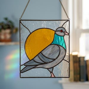 May include: A rectangular stained glass suncatcher featuring a grey pigeon with a teal neck, a bright yellow circular sun, and a small red eye. The bird is set against a textured clear glass background and hangs from a metal chain.
