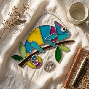 以下が含まれることがあります： 青、黄色、マゼンタの色合いのステンドグラスのカメレオンが、緑の葉が付いた茶色の枝に止まっています。アートワークはシルバーチェーンで吊り下げられています。白いマグカップ、ペン、ヴィンテージの本が背景にあります。