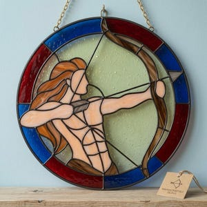 May include: A circular stained glass suncatcher depicting a figure with long hair drawing a bow and arrow. The central figure is rendered in peach and brown tones against a pale green background, framed by alternating red and blue glass segments.