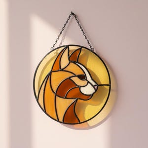 Op de afbeelding: Gebrandschilderde katten silhouet in tinten oranje, bruin en crème, tegen een gele cirkelvormige achtergrond. Het kattenprofiel is zwart omlijnd en het stuk hangt aan een metalen ketting. Een decoratieve zonnevanger.