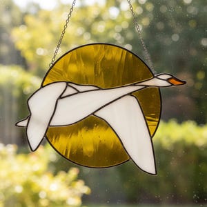 May include: A circular stained glass suncatcher featuring a white swan in flight against a translucent amber yellow background. The swan is crafted with opaque white glass pieces and black lead lines, suspended by a thin metal chain for display.