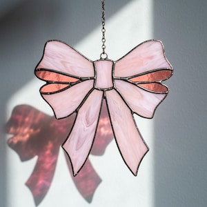 May include: A stained glass suncatcher shaped like a bow, constructed from pale pink marbled glass panels with dark metal solder lines. The bow features a central knot and two hanging tails, suspended from a thin metal chain for hanging display.