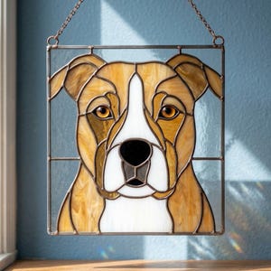 May include: A square stained glass suncatcher featuring a tan and white dog portrait with amber eyes and a black nose. The piece is framed in dark metal with a thin chain attached to the top corners for hanging, showcasing clear glass borders.