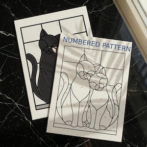 May include: Two rectangular sheets of paper featuring cat designs. One sheet shows a black cat illustration, while the other is a numbered pattern with two cats, labeled "NUMBERED PATTERN" in blue text, with black outlines and numbered sections.